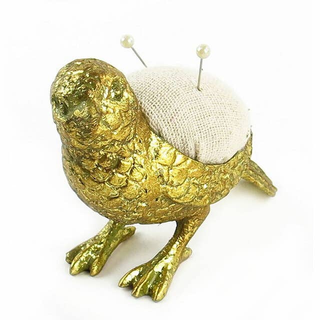 The Vagabond Vintage Bird Needle Holder features a gold, textured pewter finish. A round beige cushion with two pearl-headed pins sits on its back. The bird stands on two legs and is shown against a white background.