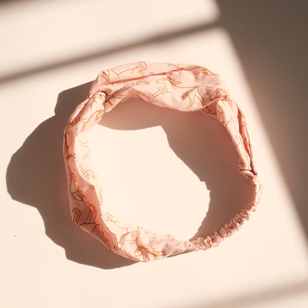 The Horses Cotton Headband by Handker Bandanas features hand screen-printed Pink Mist Horses line art on pink fabric, displayed in a loose circle on a sunlit white surface with soft shadows.