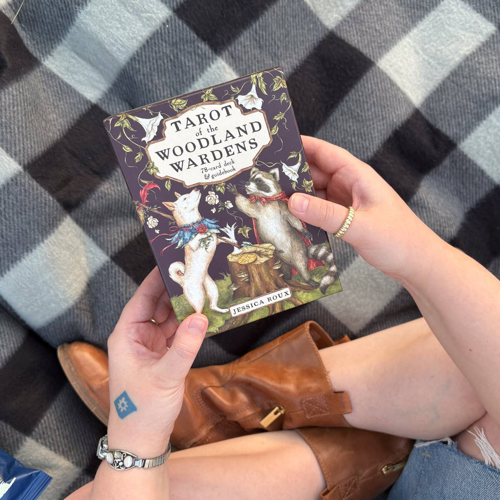A person holding a box of Simon & Schuster’s Tarot of the Woodland Wardens by Jessica Roux—an English, 224-page mixed media tarot deck—while sitting on a plaid blanket, wearing brown boots and jewelry. The box displays illustrations of animals on its cover.
