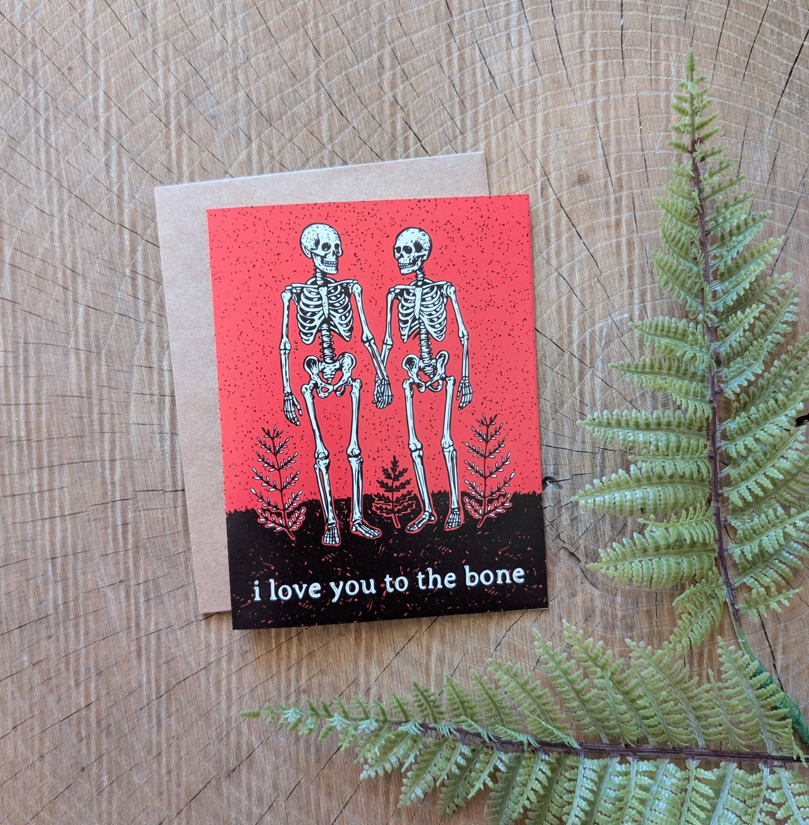 The ilikesara | art + goods greeting card, "i love you to the bone," shows two skeletons holding hands on a red background—a unique romantic card resting beside a green fern leaf.