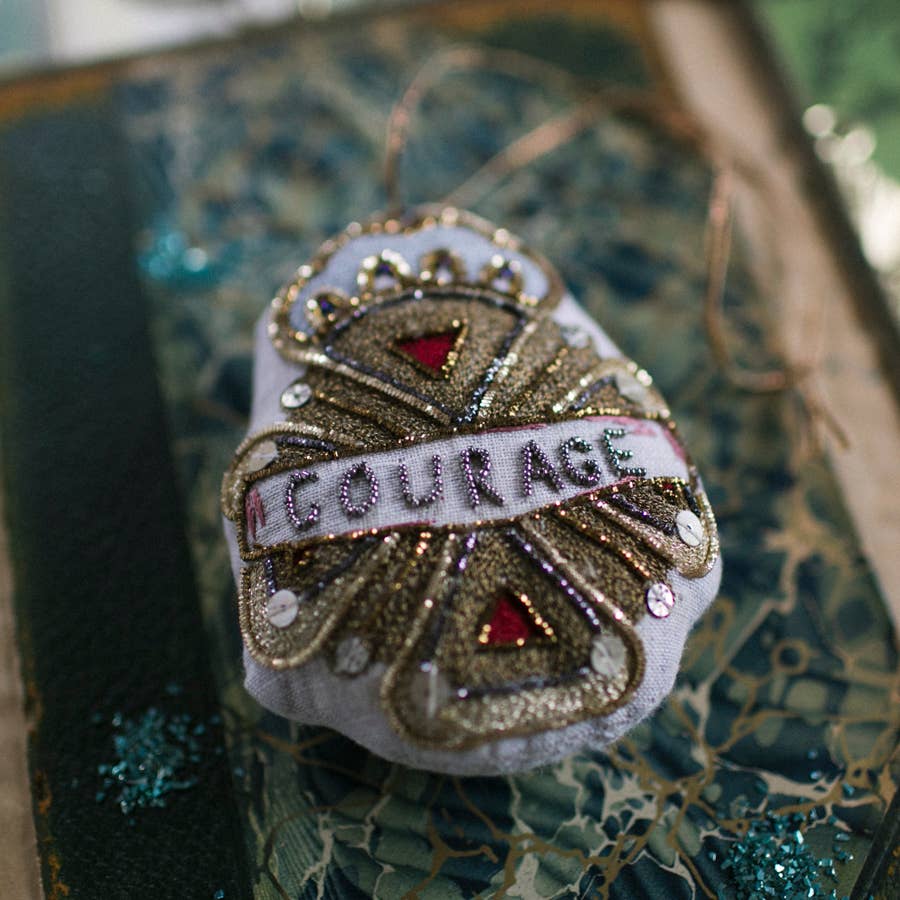 The Skippy Cotton Courage Crest Token is a cotton-filled ornament, embellished with gold, silver, and red sequins like a Red Brick Road, and embroidered with "COURAGE" at the center.