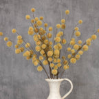 A white ceramic pitcher displays several Ragon House Faux MUSTARD GLOBE THISTLE SPRAYS with round, fluffy yellow blooms against a gray textured background.