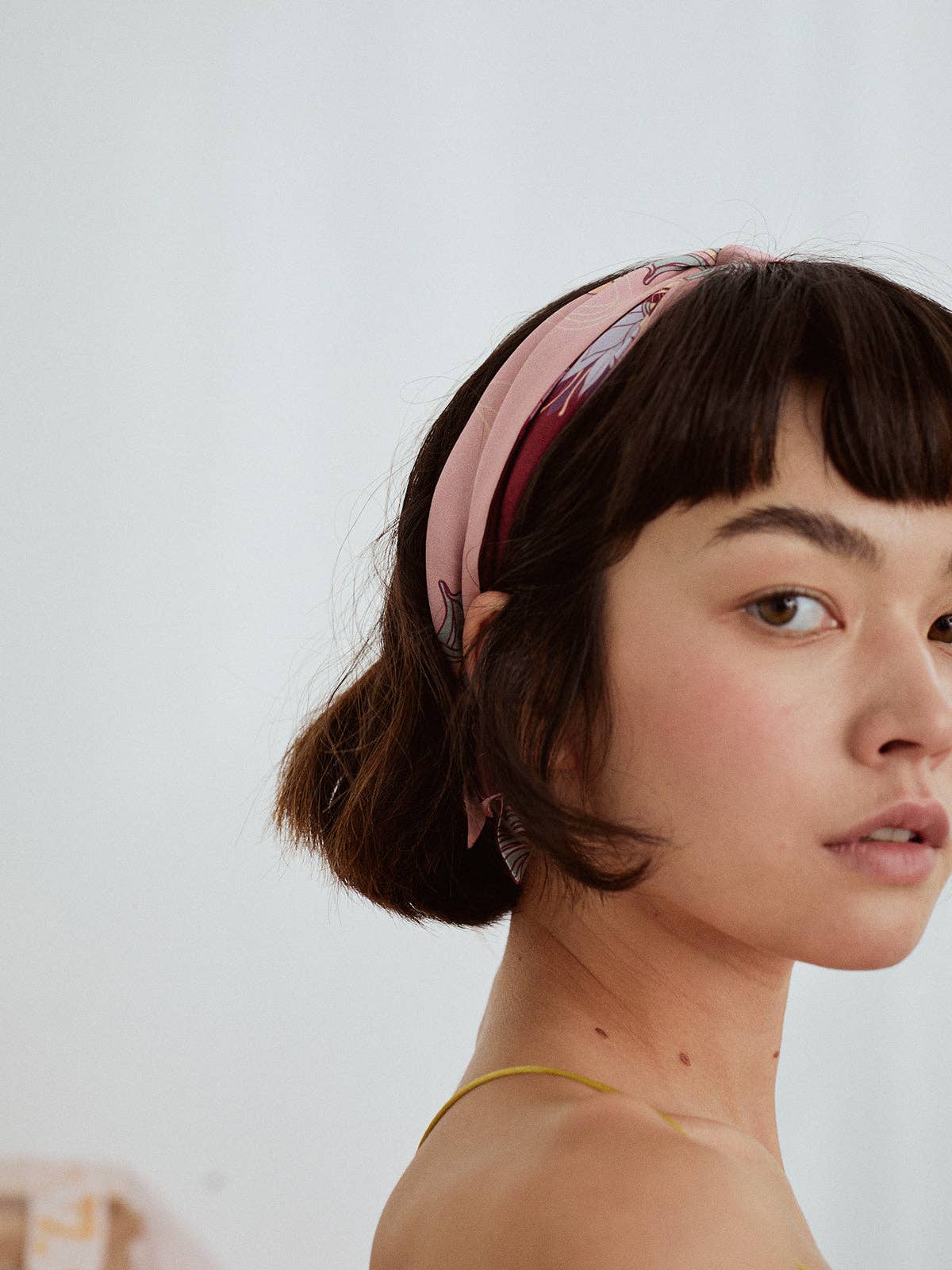 A young woman with short dark hair wears the Lost Pattern NYC Silk Skinny Scarf in Ruby Red as a chic headband, glancing over her shoulder at the camera against a plain light background.
