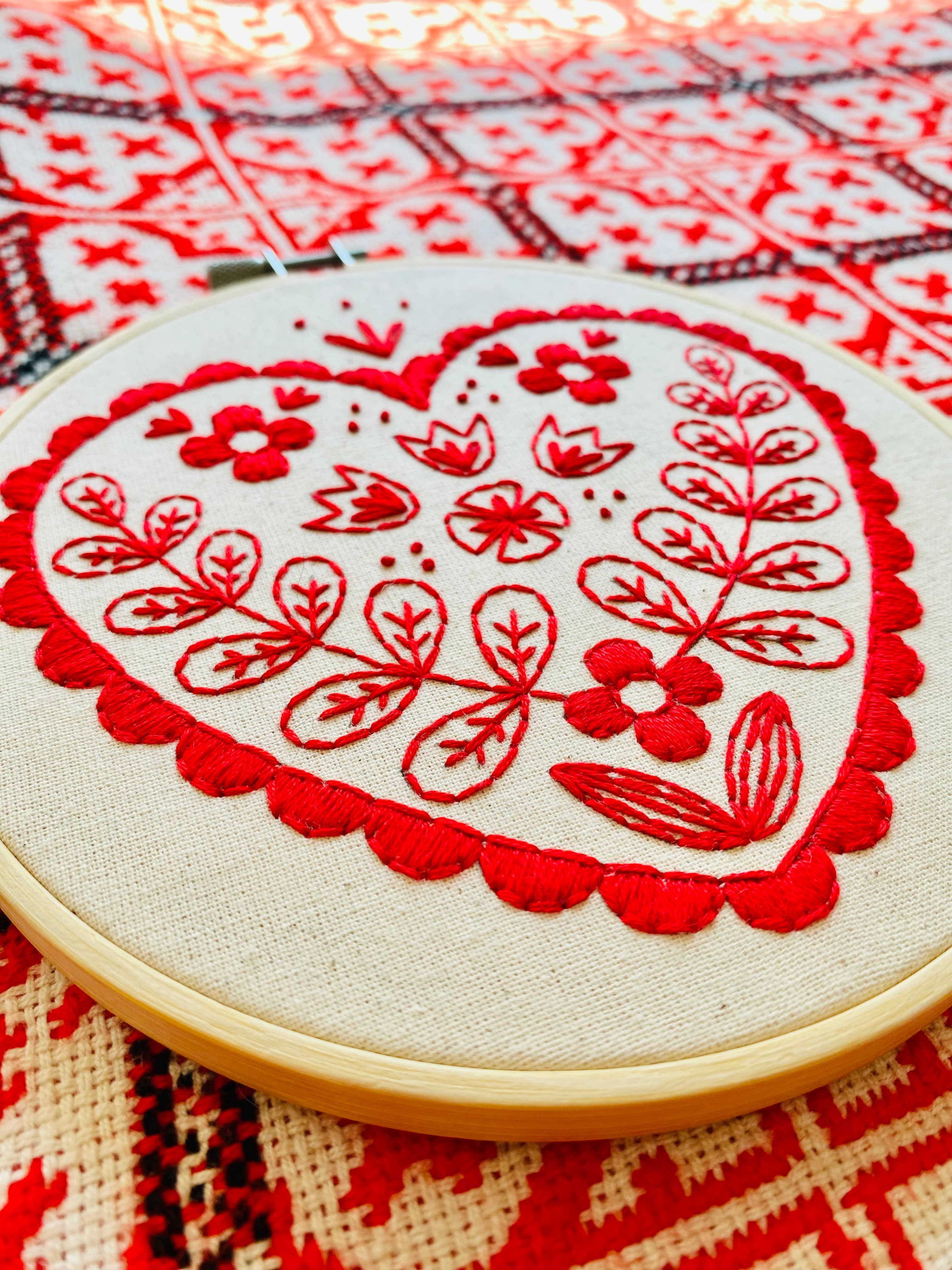 The Nordic Heart Complete Embroidery Kit by Hook, Line & Tinker features fabric with a red heart in floral and leafy motifs set against a red and white geometric background—perfect for beginner embroidery enthusiasts.