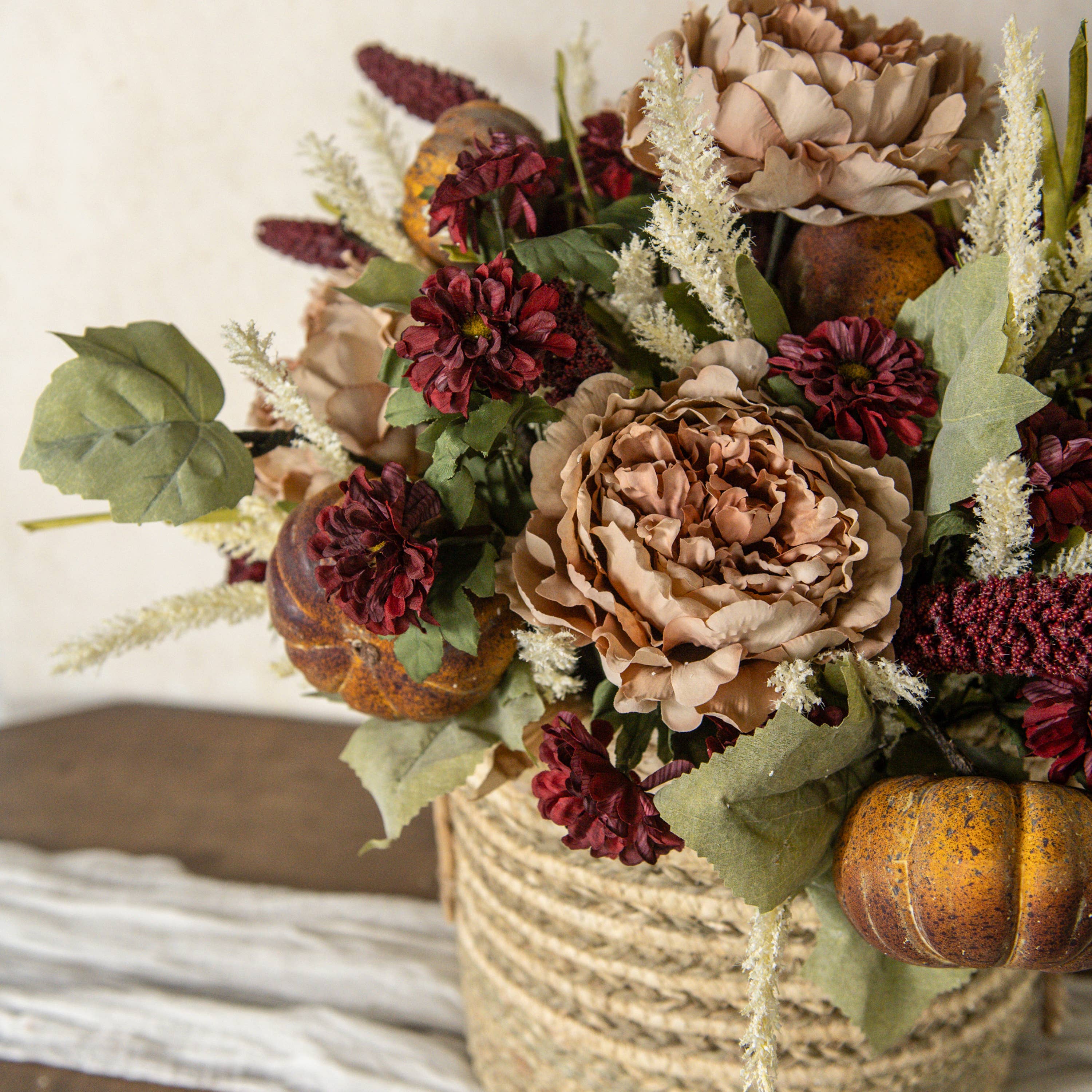 A rustic woven basket on a white cloth holds an autumn floral arrangement with Ragon House’s 31" Burgundy Sorghum Spray, beige and burgundy flowers, green leaves, dried accents, and decorative pumpkins atop a wooden surface.