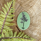 The “Iron-on patch | maidenhair fern” by ilikesara | art + goods rests on a tree stump beside a green embroidered flower patch and delicate fern leaves.