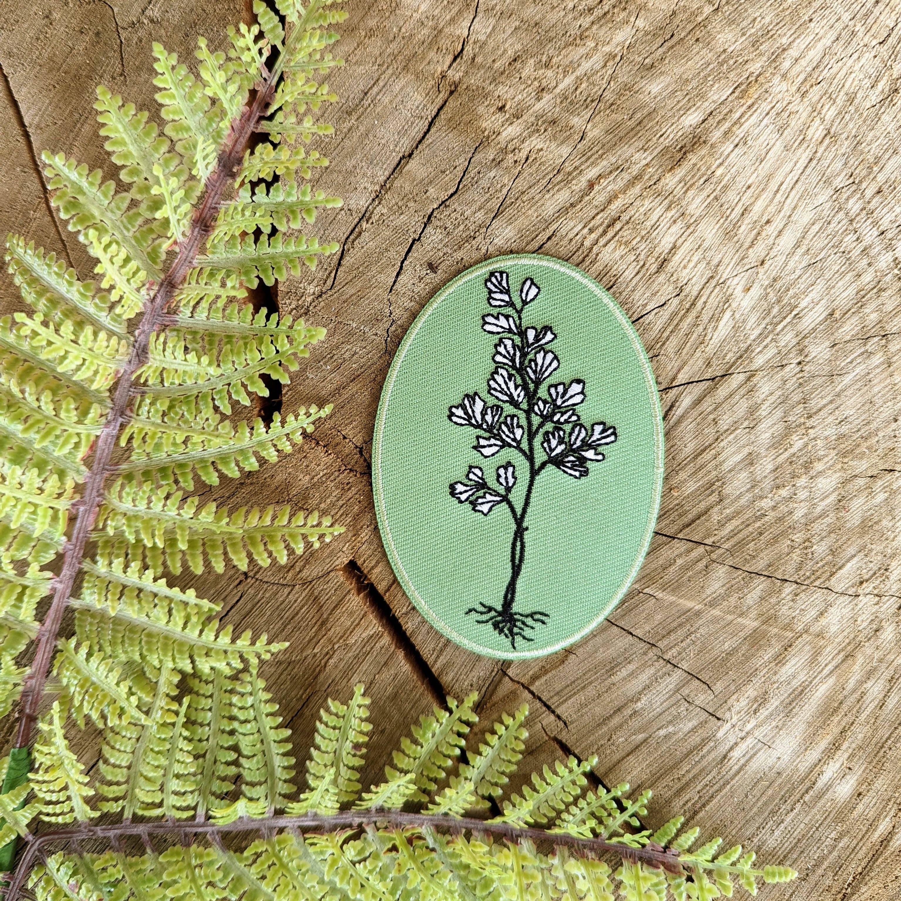 The “Iron-on patch | maidenhair fern” by ilikesara | art + goods rests on a tree stump beside a green embroidered flower patch and delicate fern leaves.