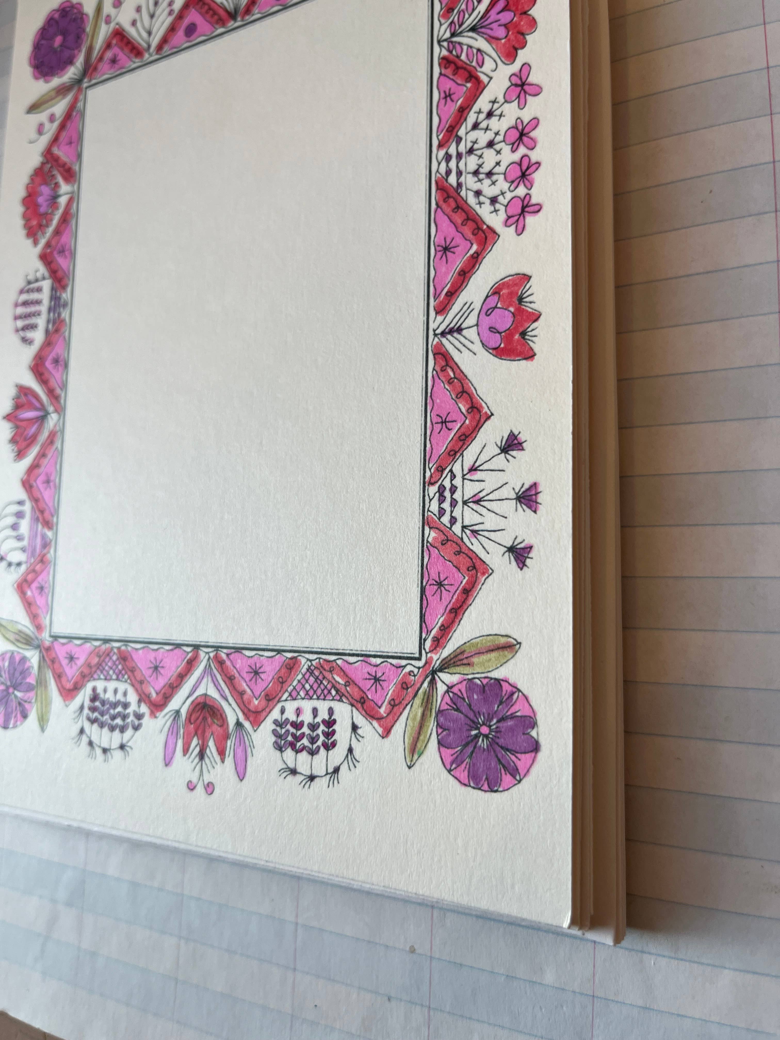 Parcel’s Colorful Folk Frame set includes 8 notebooks with blank pages framed by pink, purple, and red floral and geometric designs on quality cardstock, each notebook resting atop a lined sheet of paper.