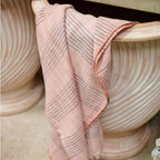 A Silica Rose Throw in shades of salmon and gray stripes by Variously is draped over the edge of a large, ribbed, cream-colored planter, with another similar planter beside it.