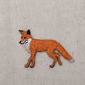 The Natural Red Fox - Walking Left - Full Body Iron on Patch by Wholesale Applique features an orange fox with a bushy tail on light beige fabric, ideal as a red fox applique or iron-on patch for your creative projects.