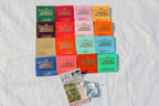 A set of colorful Fragrance Matches by H&L NOVELTIES INC, each handcrafted matchbook features a unique label and hue, arranged neatly with a match pack and silver pouch—perfect as a stylish bathroom accessory.