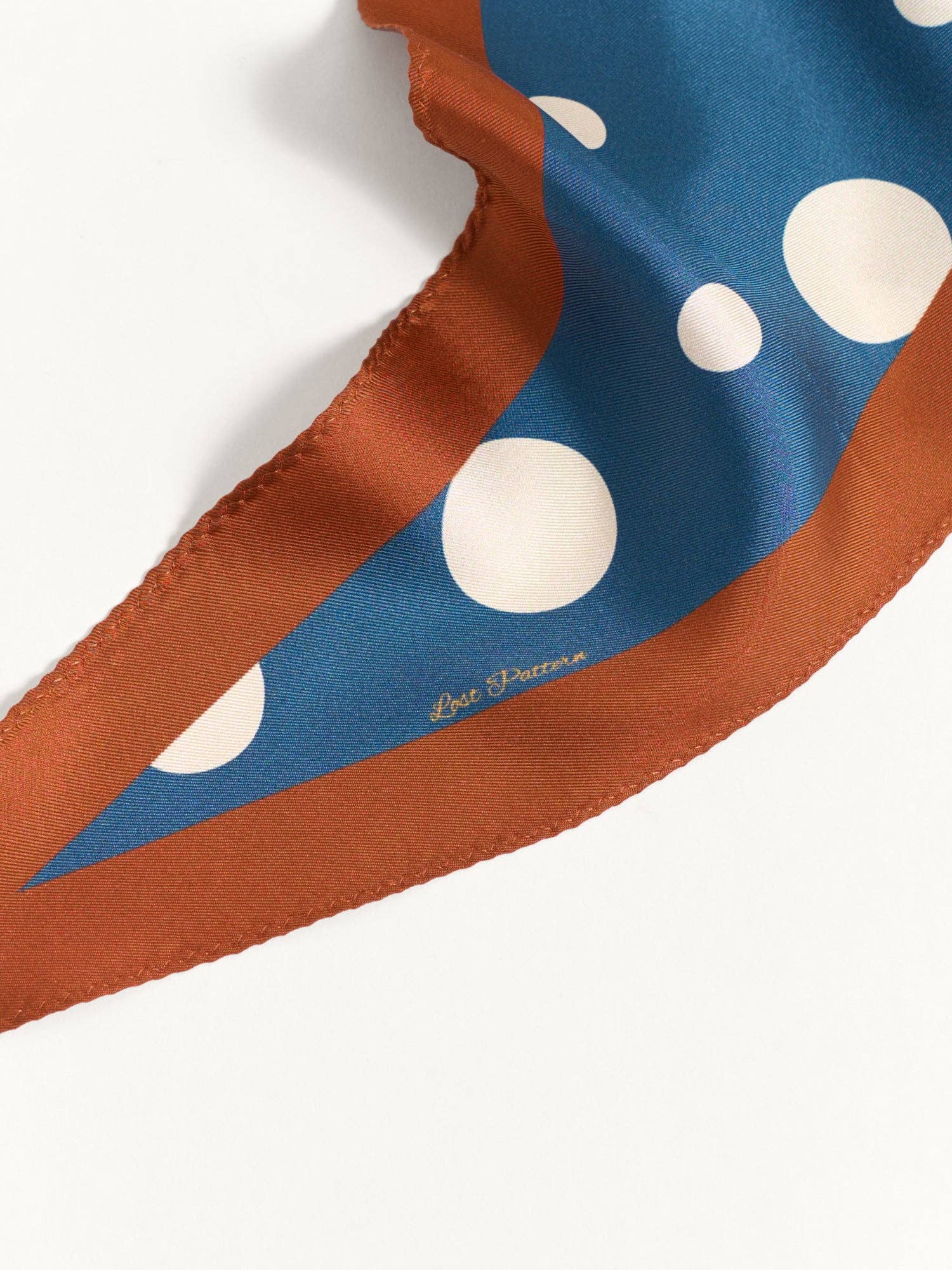 The Lost Pattern NYC Silk Polka Dot Diamond Neckerchief - Salted Caramel has a brown border, blue center with large white polka dots, and "Lost Pattern" printed near the edge. Shown partially folded on a white surface.