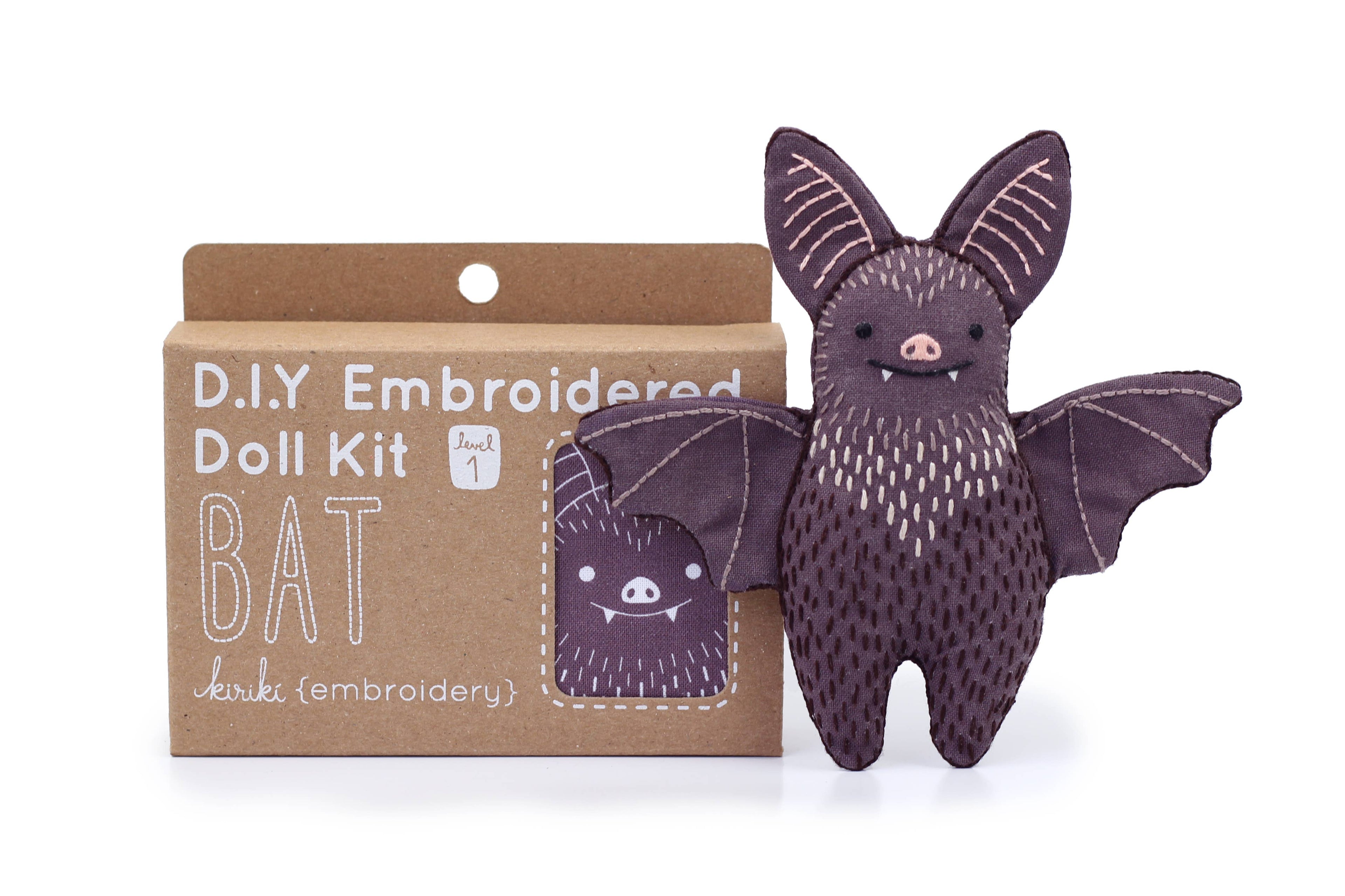 The Kiriki Press Bat - Embroidery Kit is a beginner set to make a bat plushie, featuring brown packaging with bat art and a finished purple embroidered doll shown next to it, showcasing its charming stitched details.