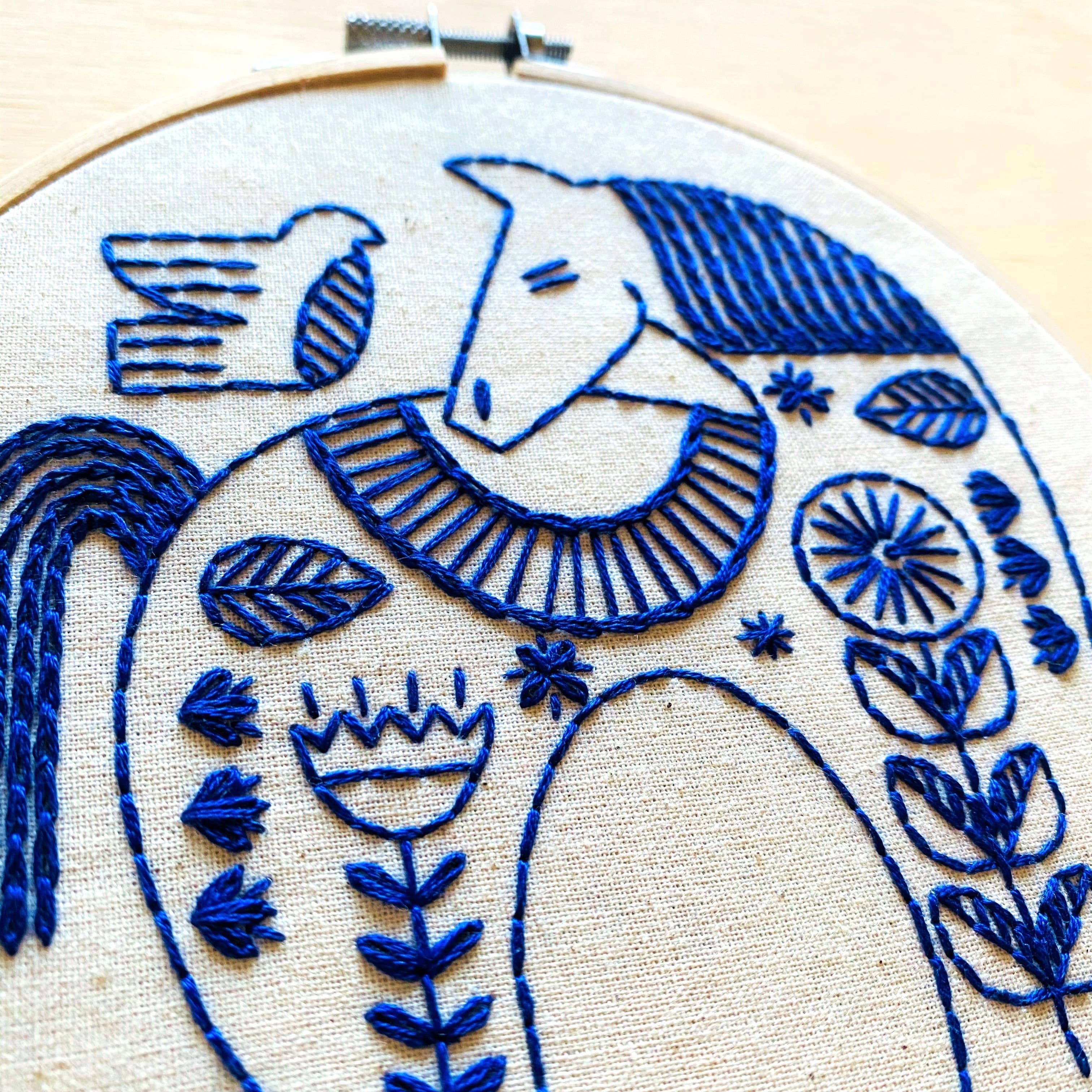 A close-up of blue embroidery on beige fabric in a hoop, featuring a stylized horse with floral, leaf, and bird motifs. The Hygge Horse Complete Embroidery Kit by Hook, Line & Tinker is perfect for craft enthusiasts.