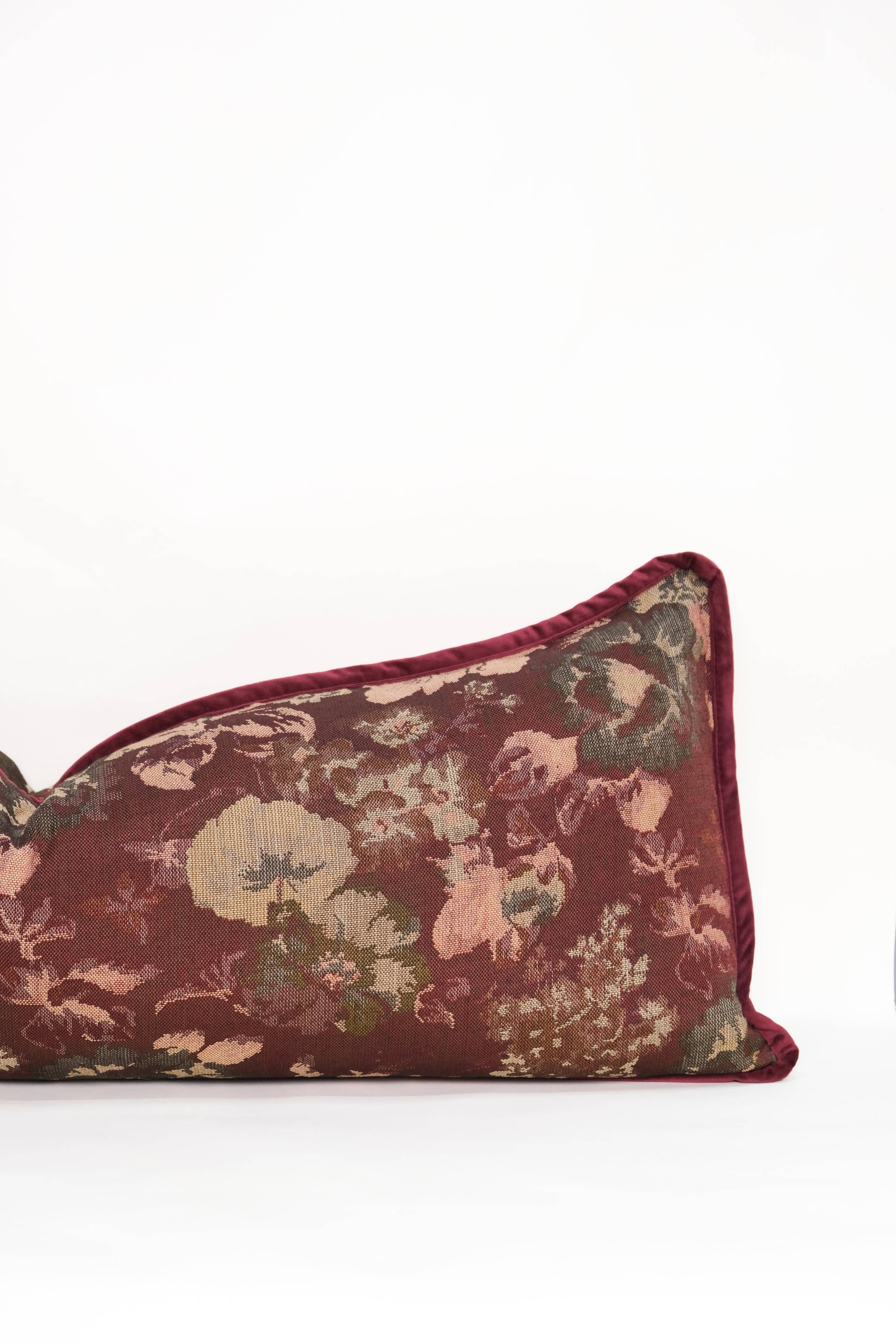 The Tapestry Long Lumbar by Twenty Third by Deanne is a rectangular maroon jacquard pillow with a floral pattern in muted greens, pinks, and beige, finished with maroon piping and shown on a plain white background.