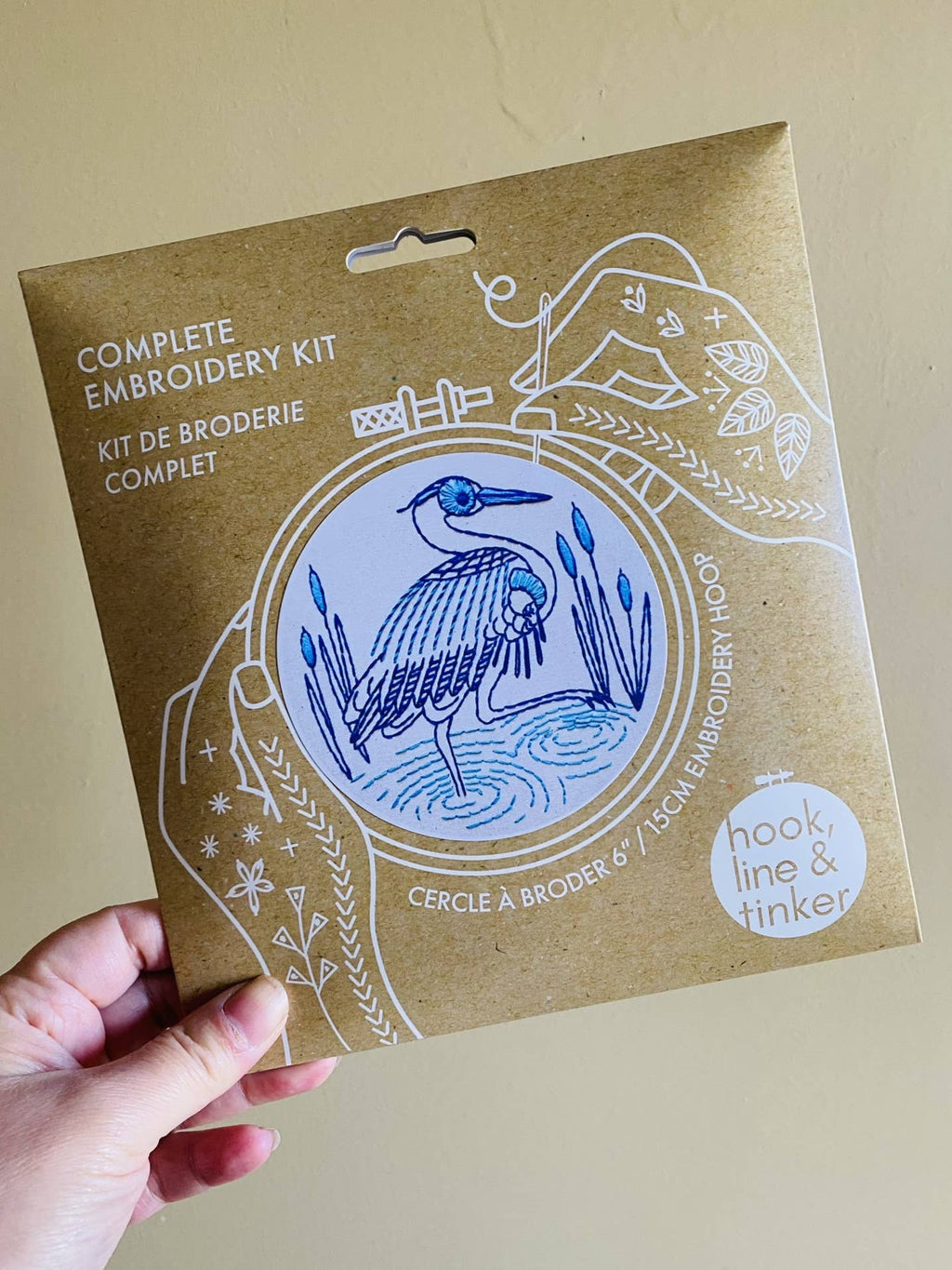 A hand holds the Hook, Line & Tinker Blue Heron Complete Embroidery Kit, featuring a blue heron design and white line art—ideal for beginner embroidery enthusiasts.