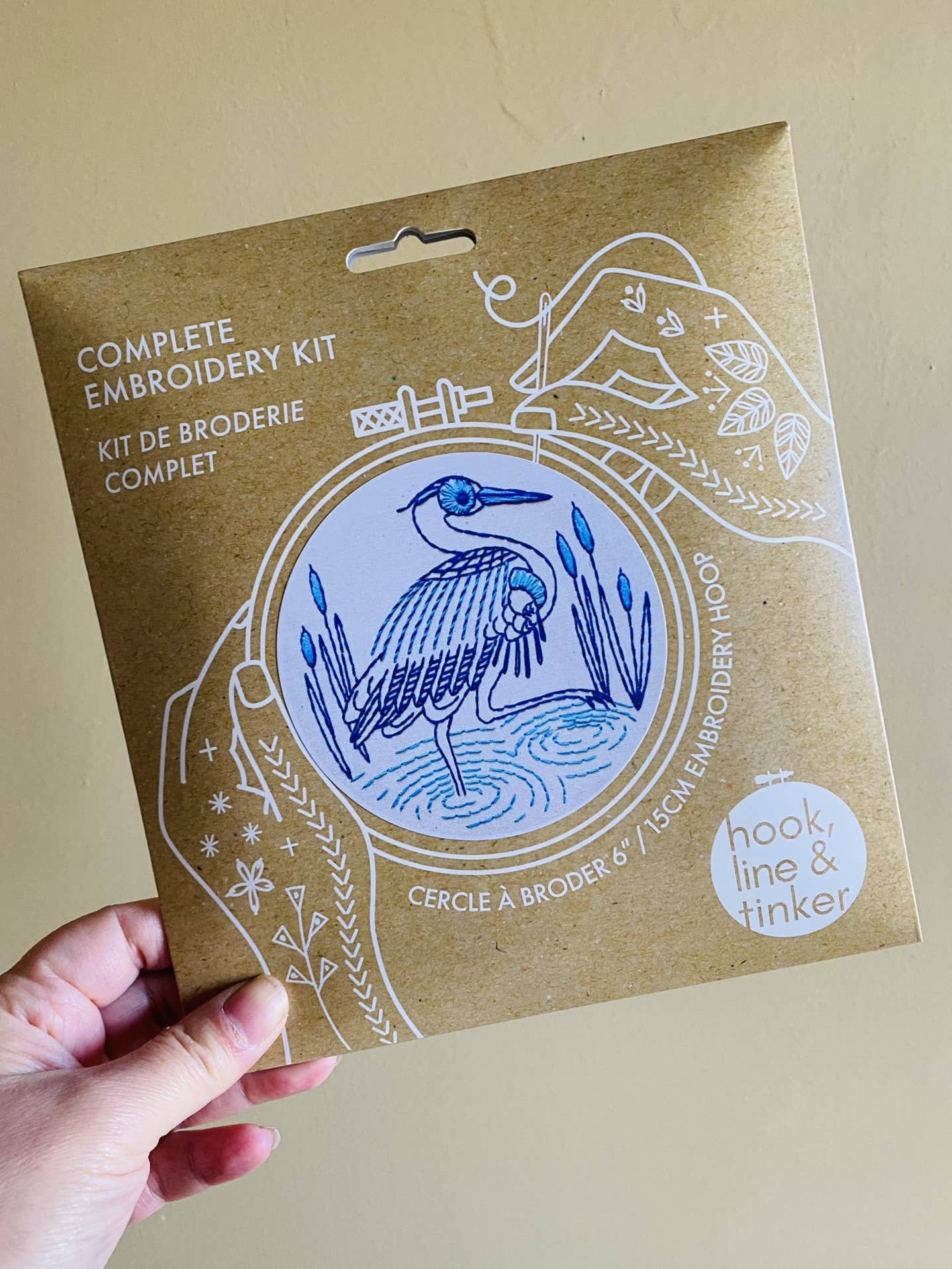 A hand holds the Hook, Line & Tinker Blue Heron Complete Embroidery Kit, featuring a blue heron design and white line art—ideal for beginner embroidery enthusiasts.