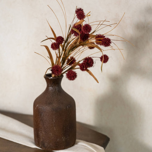 A 23" Burgundy Pom Pom Bush by Ragon House is arranged in a rustic brown ceramic vase with dried grasses, placed on a wooden surface with a light cloth against a pale, textured wall.