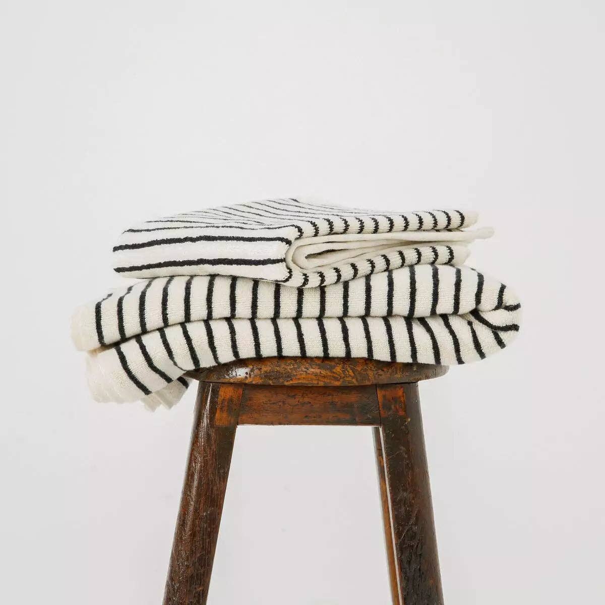 Two Turkish Cotton Striped Terry Bath Sheets by The Loomia, featuring black horizontal stripes, are neatly folded and stacked on a wooden stool against a plain white background.