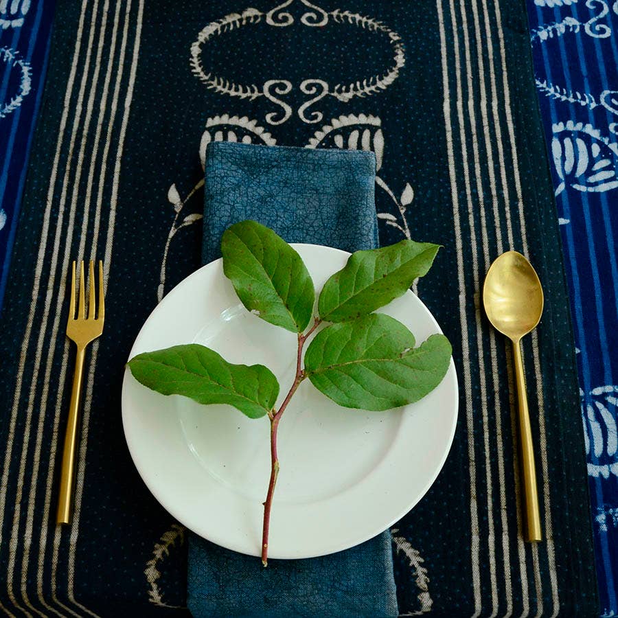 A white plate with a leafy green branch rests on Ichcha's Deepest Navy & White Table Linens Cotton Table Runner - Danya, with a gold fork left, gold spoon right, and a dark blue napkin beneath the plate.