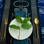 A white plate with a leafy green branch rests on Ichcha's Deepest Navy & White Table Linens Cotton Table Runner - Danya, with a gold fork left, gold spoon right, and a dark blue napkin beneath the plate.