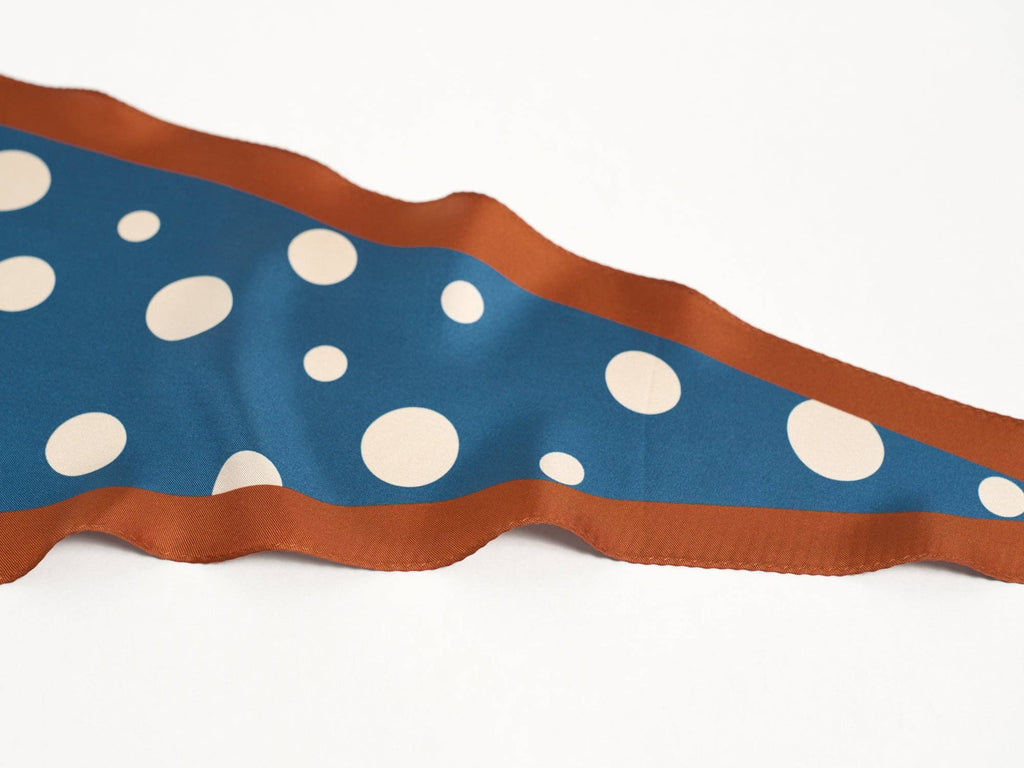 The Lost Pattern NYC Silk Polka Dot Diamond Neckerchief in Salted Caramel features blue mulberry silk with large and small white polka dots, a brown border, and a wavy triangular shape on a plain white background.