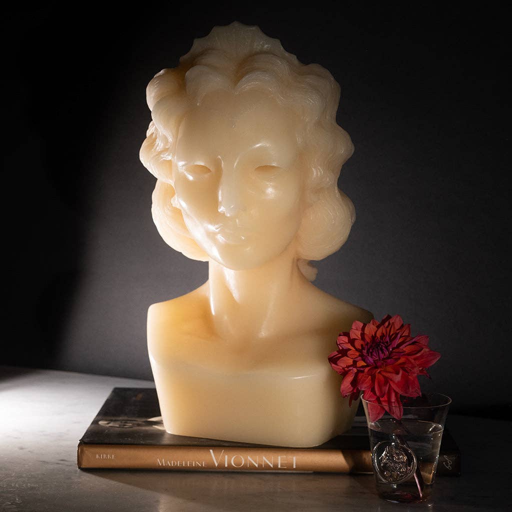 The limited edition WEEPING BUST candle by Heretic Parfum, cream-colored and shaped like a woman’s bust with wavy hair, sits on a book titled Vionnet beside a glass with a red flower, all against a dark background.