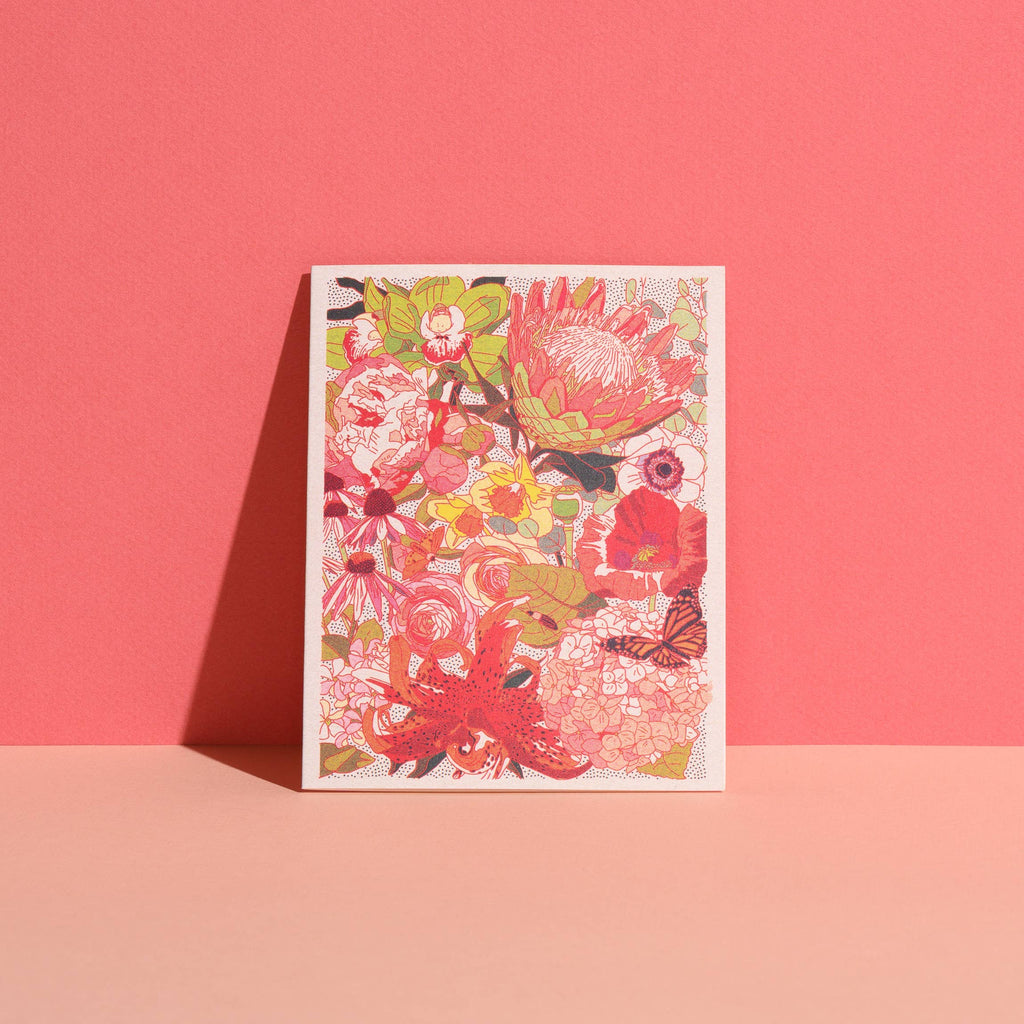 The Wild Florals greeting card by Red Cap Cards features a vibrant floral design in red, pink, yellow, and green tones. Printed in the USA on recycled paper, it stands upright against a two-tone pink background.