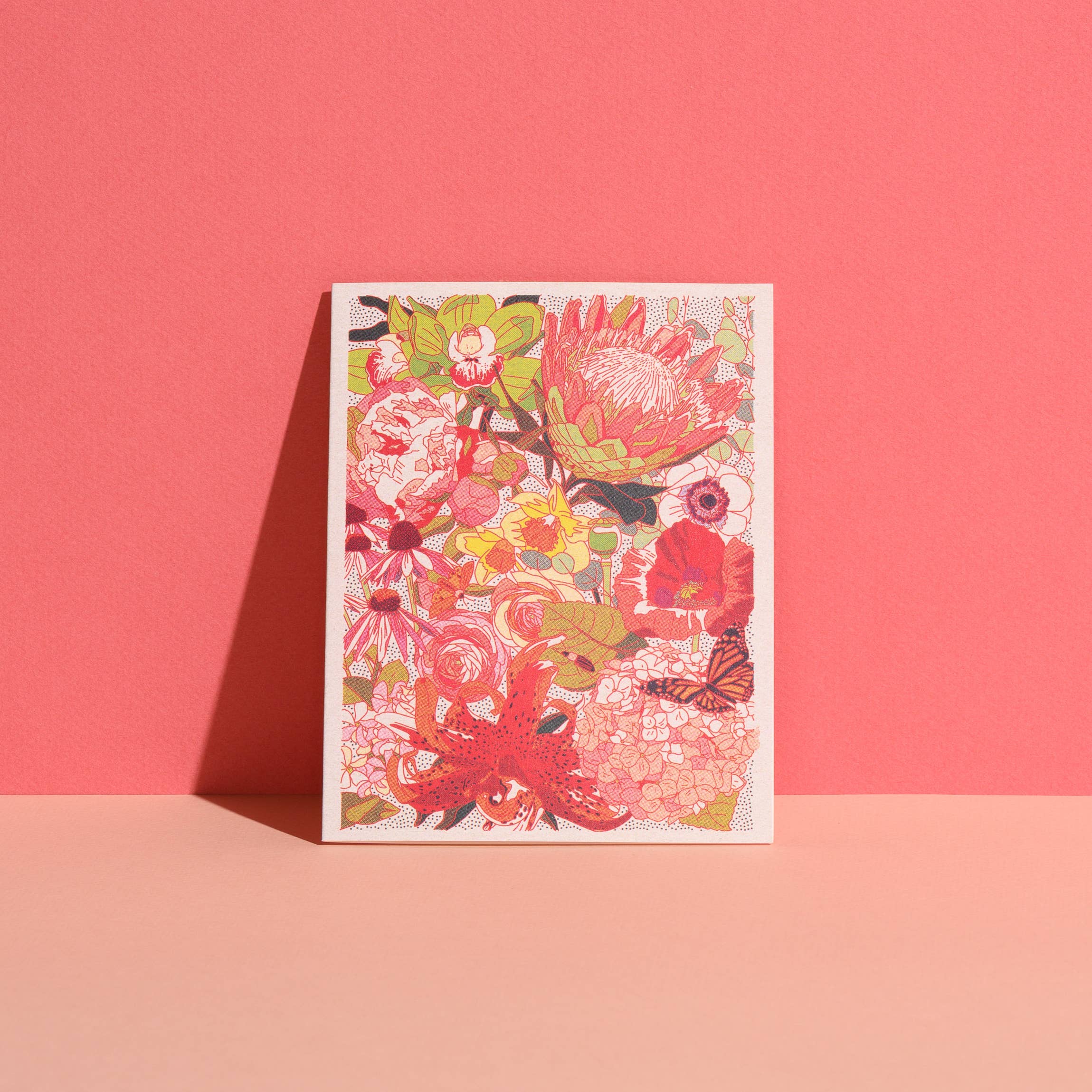 The Wild Florals greeting card by Red Cap Cards features a vibrant floral design in red, pink, yellow, and green tones. Printed in the USA on recycled paper, it stands upright against a two-tone pink background.
