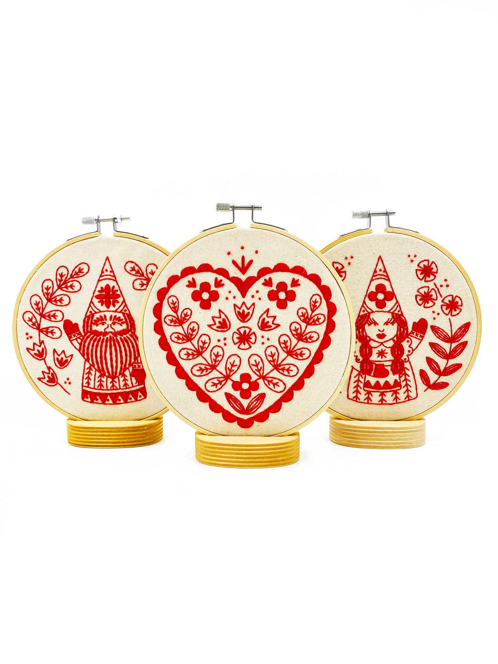 The Hook, Line & Tinker Nordic Heart Complete Embroidery Kit includes three hoops: a central hoop with a floral heart and two side hoops with gnomes among plants, all displayed on round wooden stands—perfect for beginners.