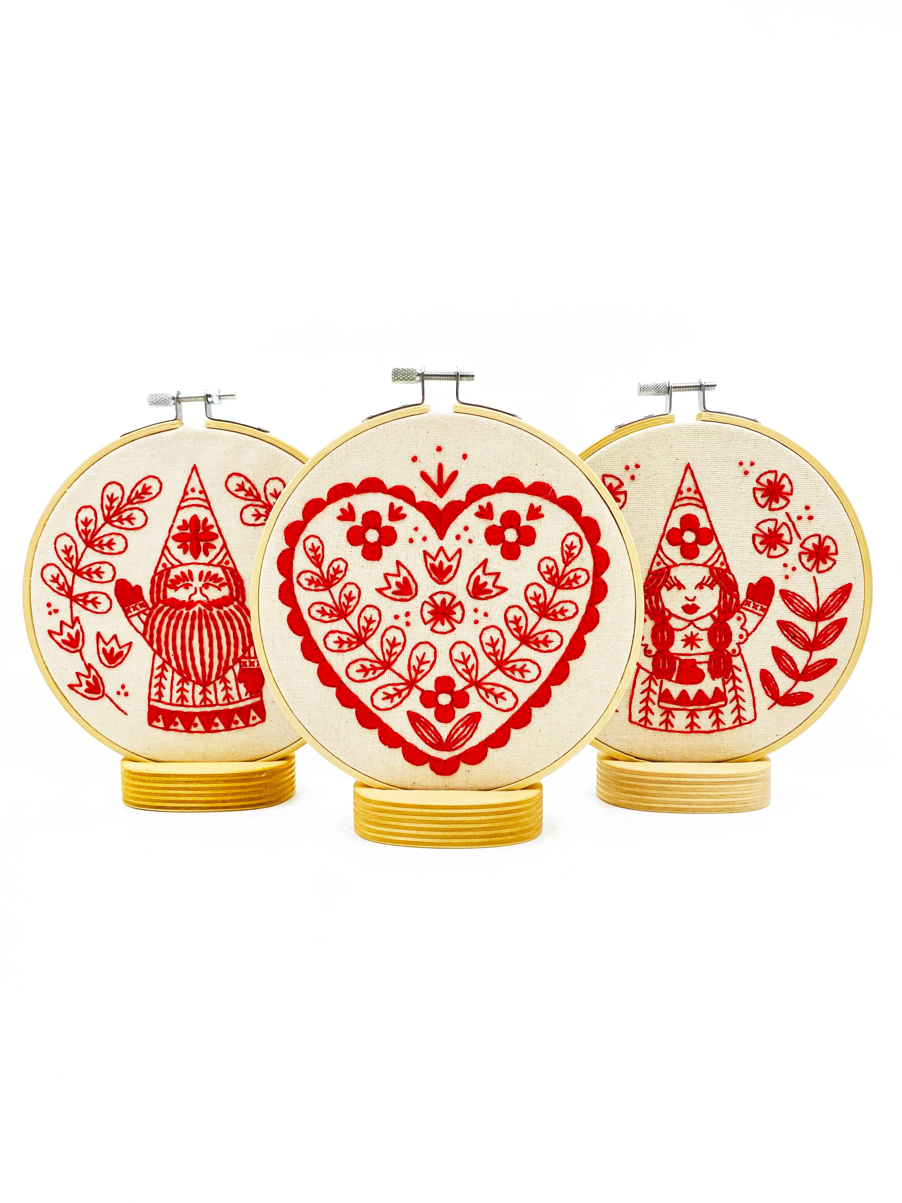 The Hook, Line & Tinker Nordic Heart Complete Embroidery Kit includes three hoops: a central hoop with a floral heart and two side hoops with gnomes among plants, all displayed on round wooden stands—perfect for beginners.