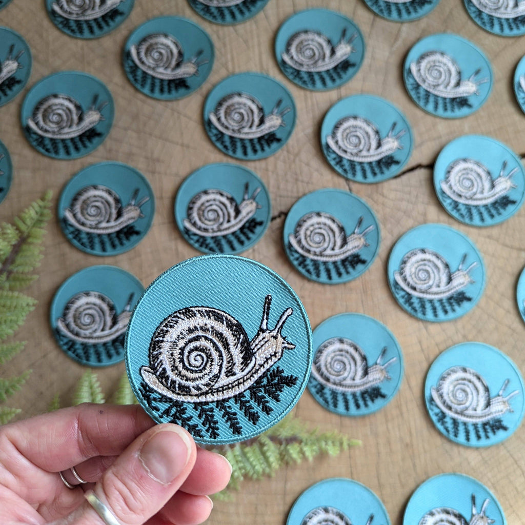 A hand holds an ilikesara | art + goods 2.5" round teal iron-on patch featuring a snail with a fern on a blue background. Multiple identical patches are spread on wood with fern leaves partly visible nearby. No backer card or packaging shown.