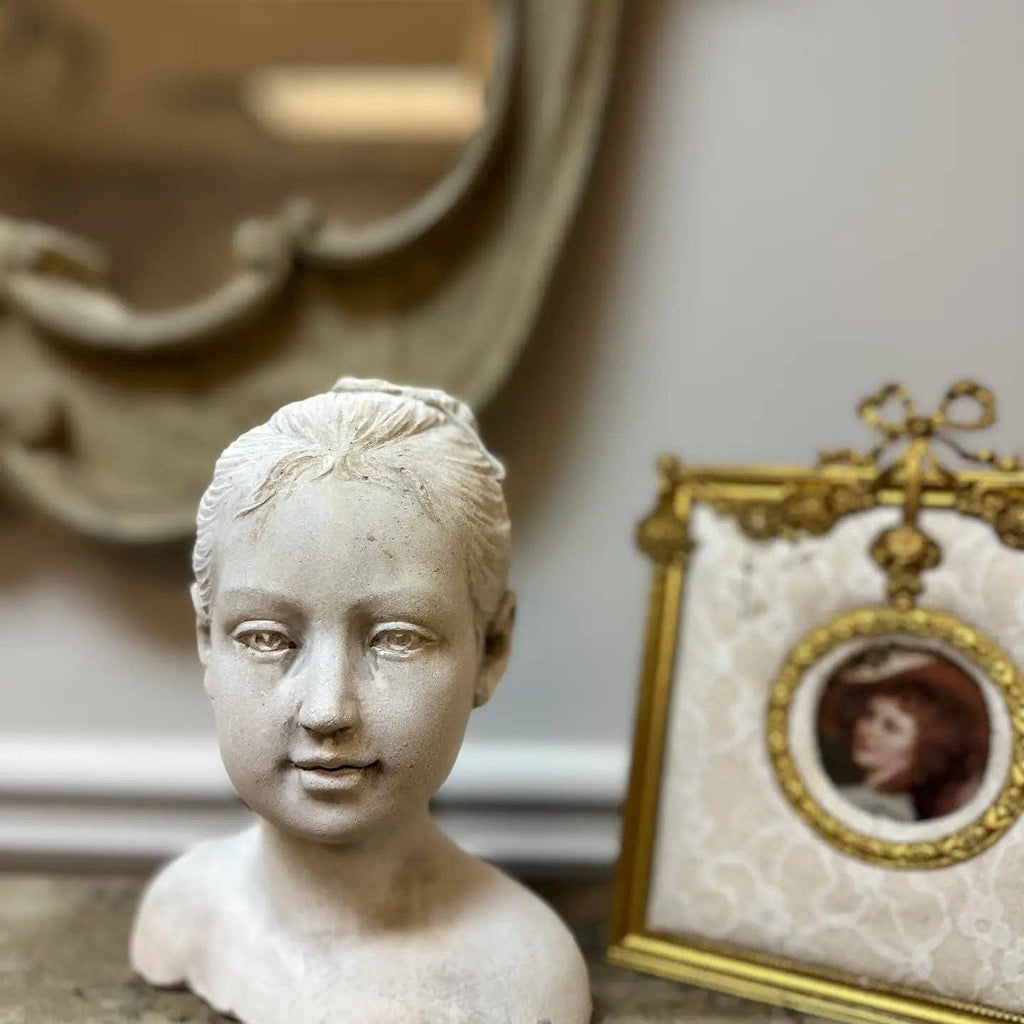 A Vagabond Vintage Cement Girl Statue is displayed on a surface beside a framed vintage portrait of a woman in a hat, with a decorative mirror hanging in the blurred background.
