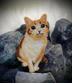 The Orange Tabby Cat embroidered iron-on patch by Wholesale Applique features realistic green eyes and sits among rough gemstones on a soft white background.