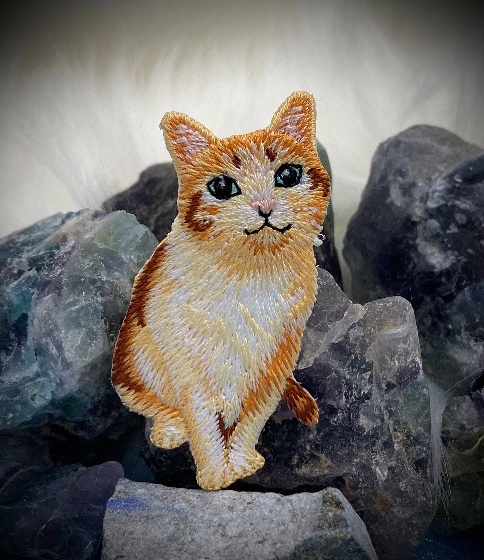 The Orange Tabby Cat embroidered iron-on patch by Wholesale Applique features realistic green eyes and sits among rough gemstones on a soft white background.