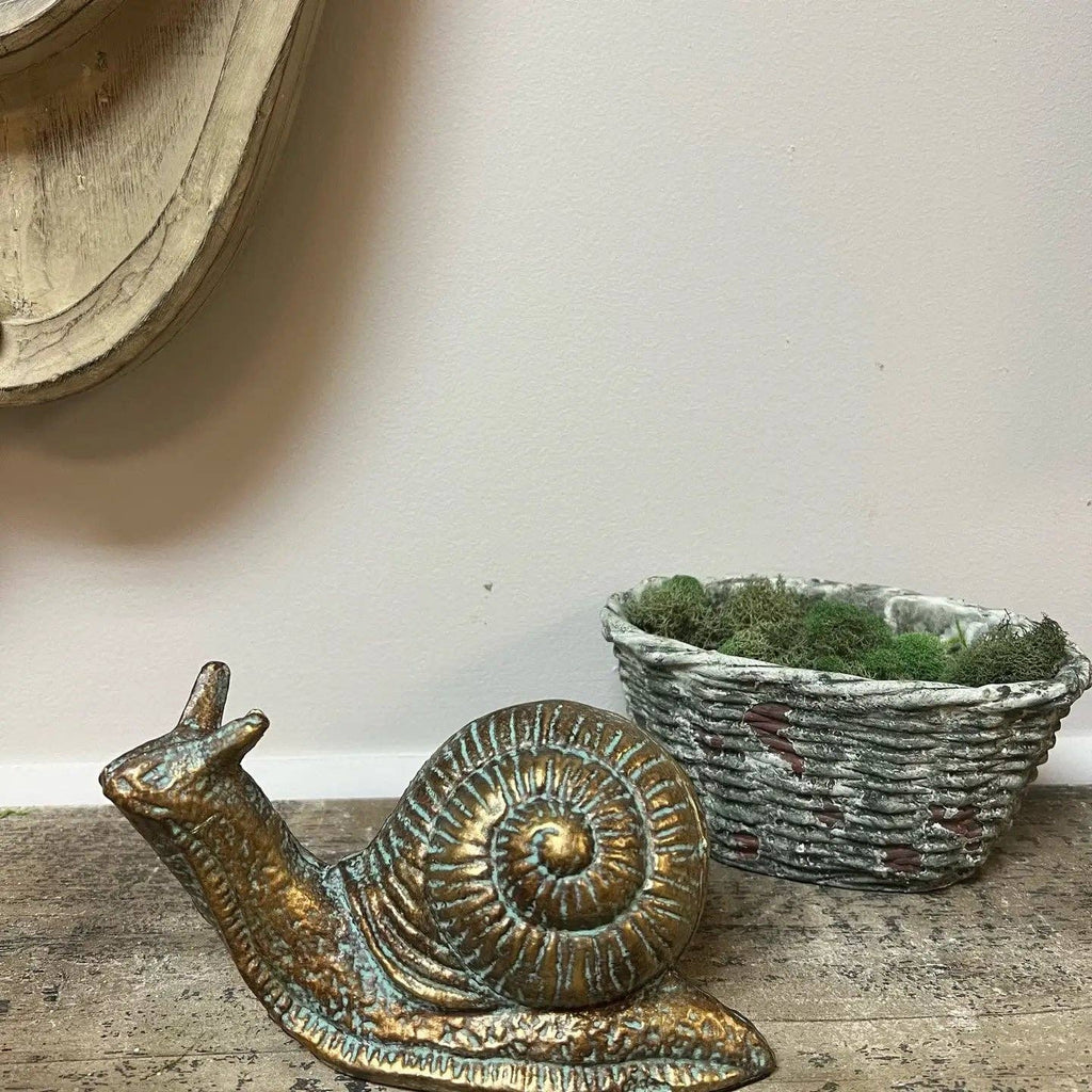 The Vagabond Vintage CAST IRON SNAIL STATUE in gold adds a charming accent to your home decor, displayed on a wooden surface next to a white woven basket with moss against a light beige wall.