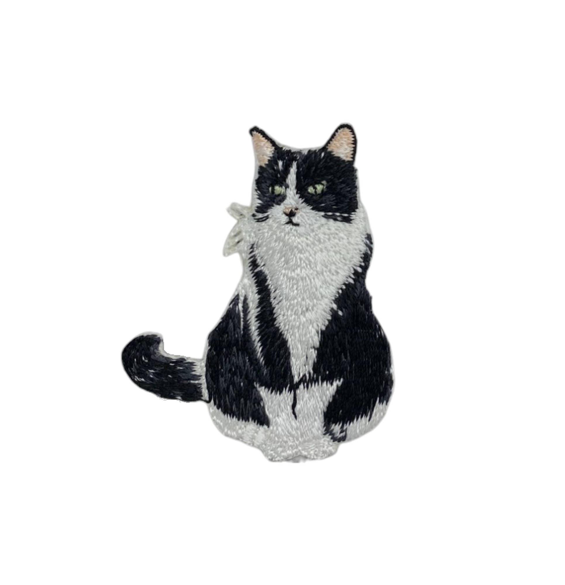 The Tuxedo Cat Iron on Patch by Wholesale Applique features a detailed black and white sitting kitten with green eyes, curled tail, and textured fur, against a white background—perfect for pet lovers.