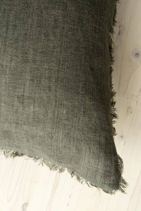 A close-up of the Indaba Trading 24x24 Lina Linen Pillow Avocado: Cover with Filler, featuring dark gray textured linen and fringed edges, crafted from sustainable materials and set on a light wooden surface.