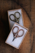 NNK Press Mini Embroidery Scissors in Antique Gold, featuring compact stainless steel blades, are displayed in small white-padded cardboard boxes on a rustic wooden surface.