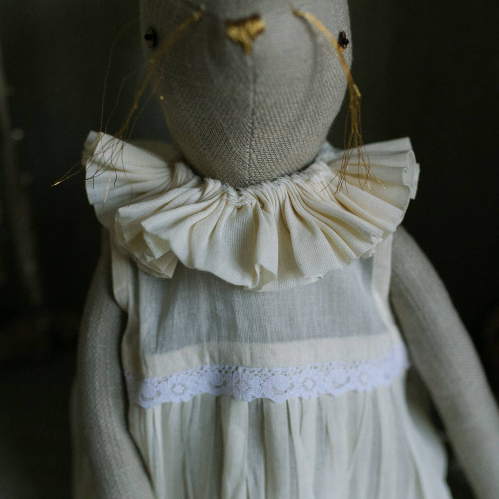 The Skippy Cotton Ruffle Bun Ragdoll is a handmade linen rabbit doll with long thread whiskers, hand-stitched details, and a ruffled cream cotton dress with lace trim, set against a neutral background.