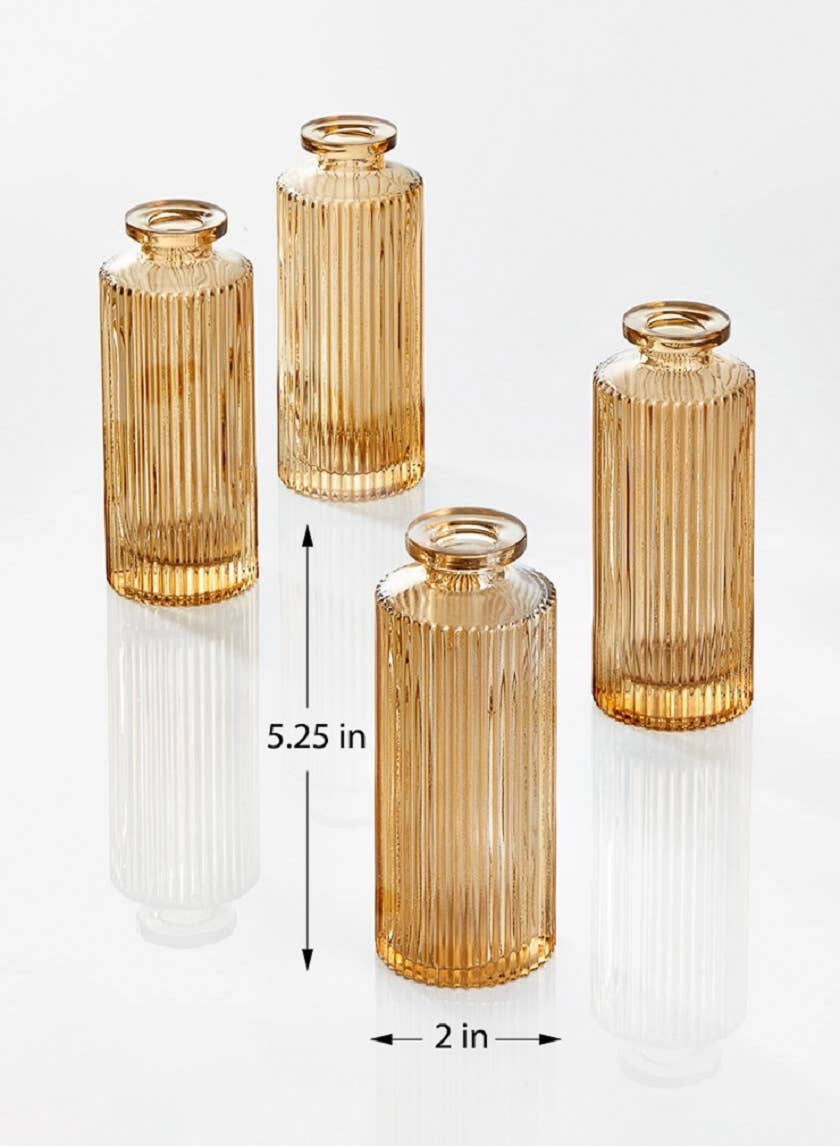 Four Jamali Garden Amber Pleated Glass Bottle Bud Vases, each 5.25" tall and 2" wide, are arranged on a white surface. The vases feature a pleated design with subtle reflections visible beneath them.