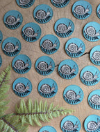 Several 2.5" round teal iron-on snail with fern patches by ilikesara | art + goods are arranged on cracked wood, accented with green fern leaves in the corner. No backer card or packaging included.