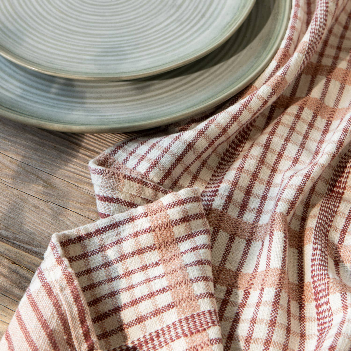 Two ceramic plates with a light green glaze and circular patterns rest on a wooden surface beside the Danica Heirloom Bisque Finley Check Dishtowels Set of 2 in classic red and white.