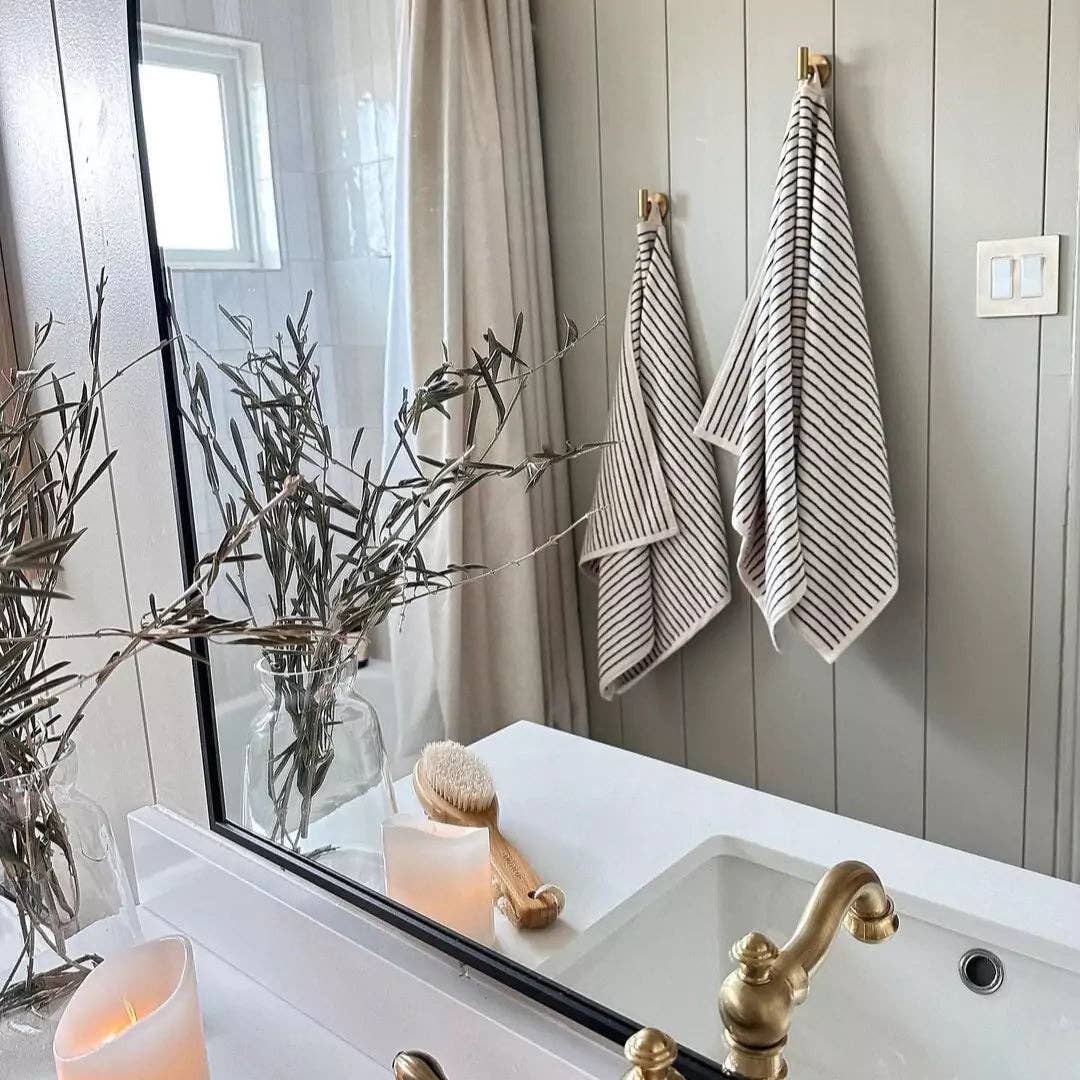 A bathroom features a gold faucet, white sink, candle, wooden brush, glass vase with dried branches, and two The Loomia Turkish Cotton Striped Terry Bath Towels hanging near a light switch and shower curtain.