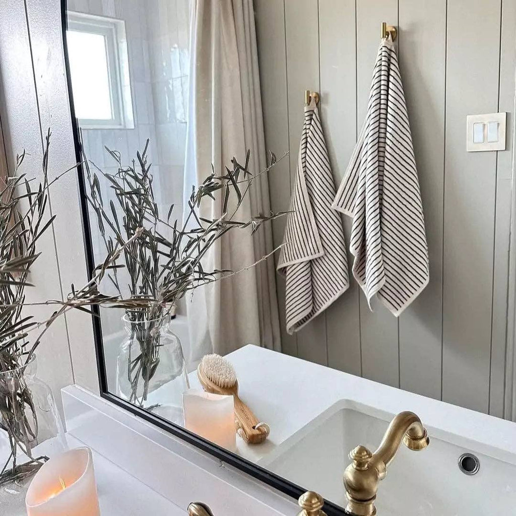 A bathroom features a gold faucet, white sink, candles, and two The Loomia Turkish Cotton Striped Terry Towel Bath Sheets hanging on the wall. A jar with branches, a wooden brush on the counter, and the mirror reflects a shower curtain and window.