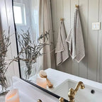 A bathroom features a gold faucet, white sink, candles, and two The Loomia Turkish Cotton Striped Terry Towel Bath Sheets hanging on the wall. A jar with branches, a wooden brush on the counter, and the mirror reflects a shower curtain and window.
