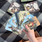 A hand holds several illustrated cards from the Tarot of the Woodland Wardens by Jessica Roux (Simon & Schuster), a mixed media deck featuring woodland animals, displayed on a checkered blanket with its box and nature-inspired tarot cards in the background.