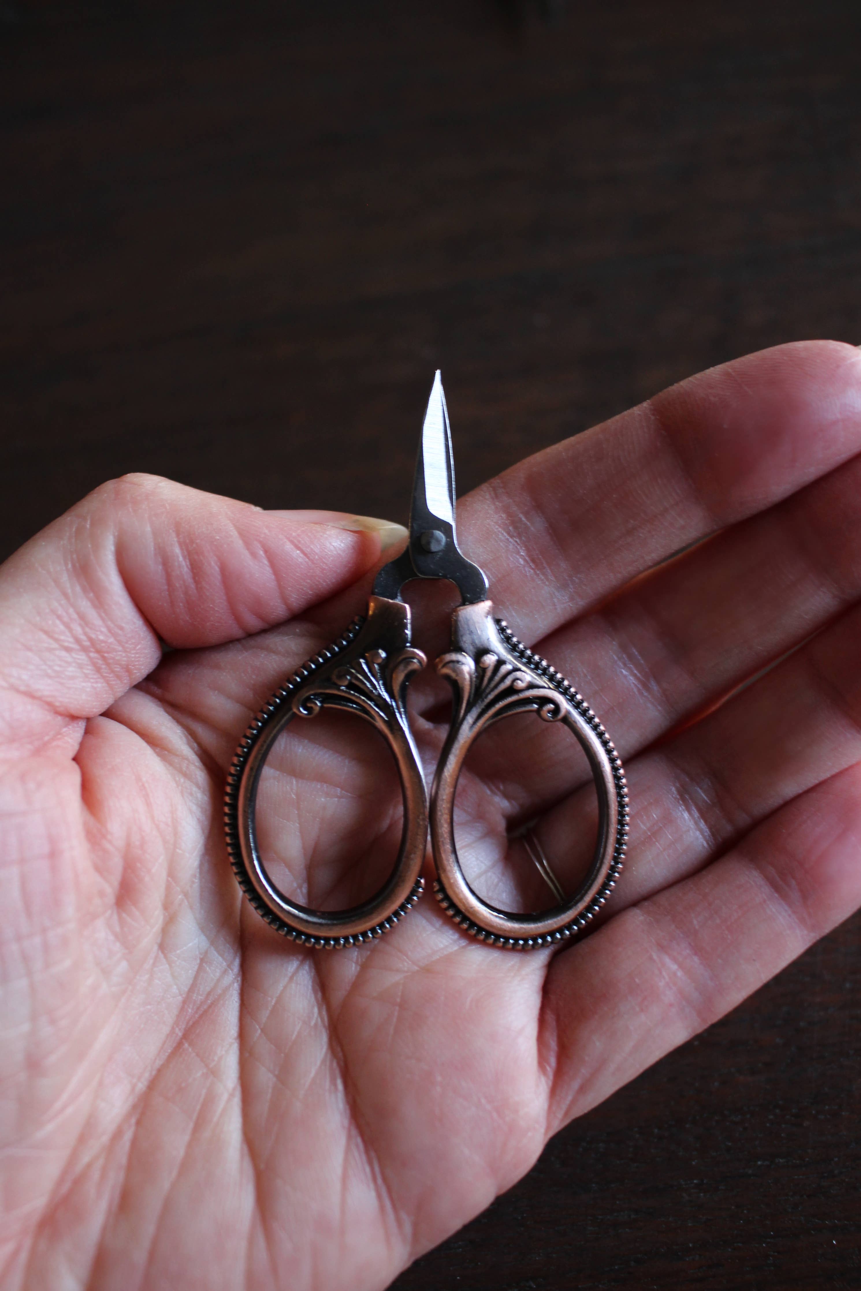 A hand holds NNK Press Mini Embroidery Scissors in Antique Gold, featuring ornate handles and sharp stainless steel blades—perfect compact sewing snips for your travel notions kit.