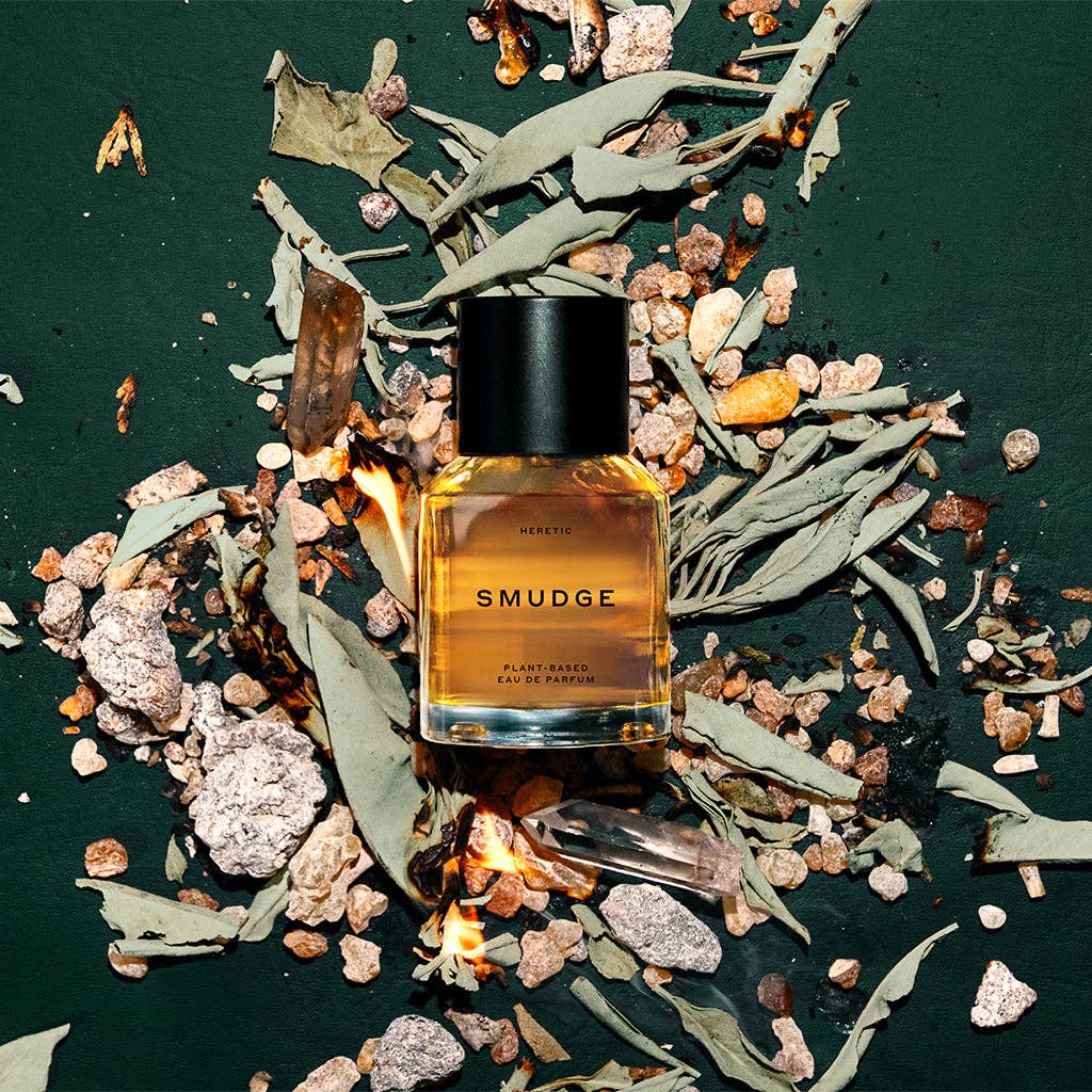 A 15ml bottle of Heretic Parfum's SMUDGE rests on a dark green surface amid dried sage, crystals, stones, and small flames—its scent blending frankincense and tonka bean.