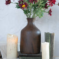 A 9.75" BROWN & RUST TONE VASE by Ragon House, filled with red and yellow flowers, sits atop vintage books between two lit white pillar candles against a textured white background.